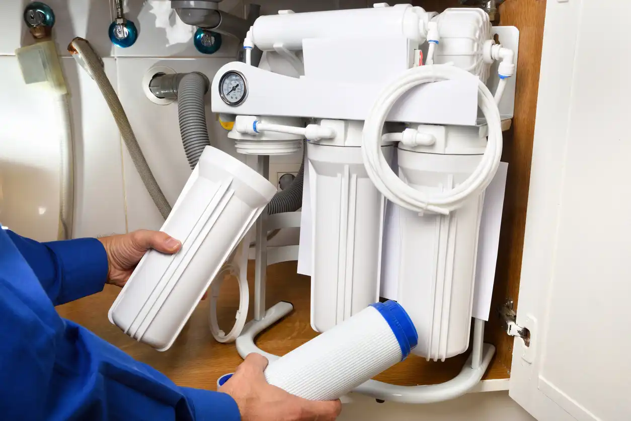 A person's hands hold a replacement water filter cartridge and its housing, with a multi-stage water filter system installed under a sink in the background