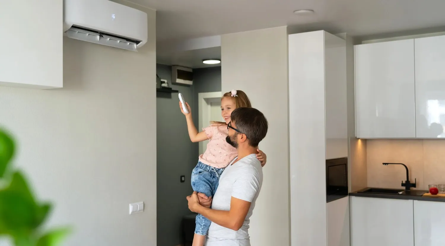Father holding daughter with remote control, adjusting ductless mini-split air conditioner in modern kitchen, emphasizing indoor air quality and HVAC efficiency