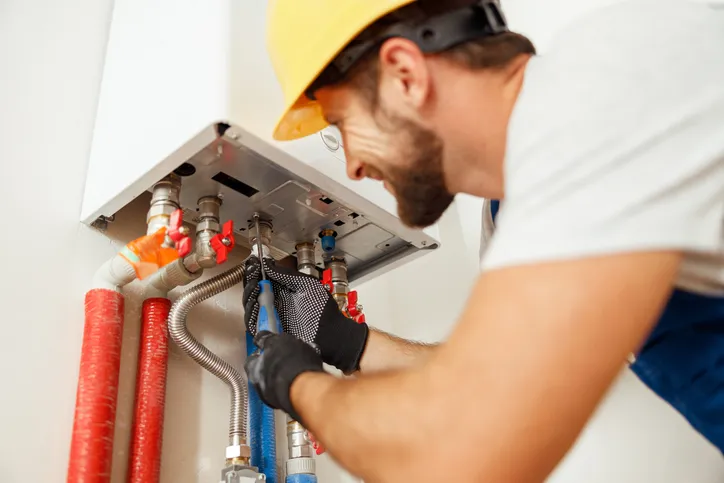 A bearded male plumber in a hard hat and gloves is using a wrench to work on a tankless water heater