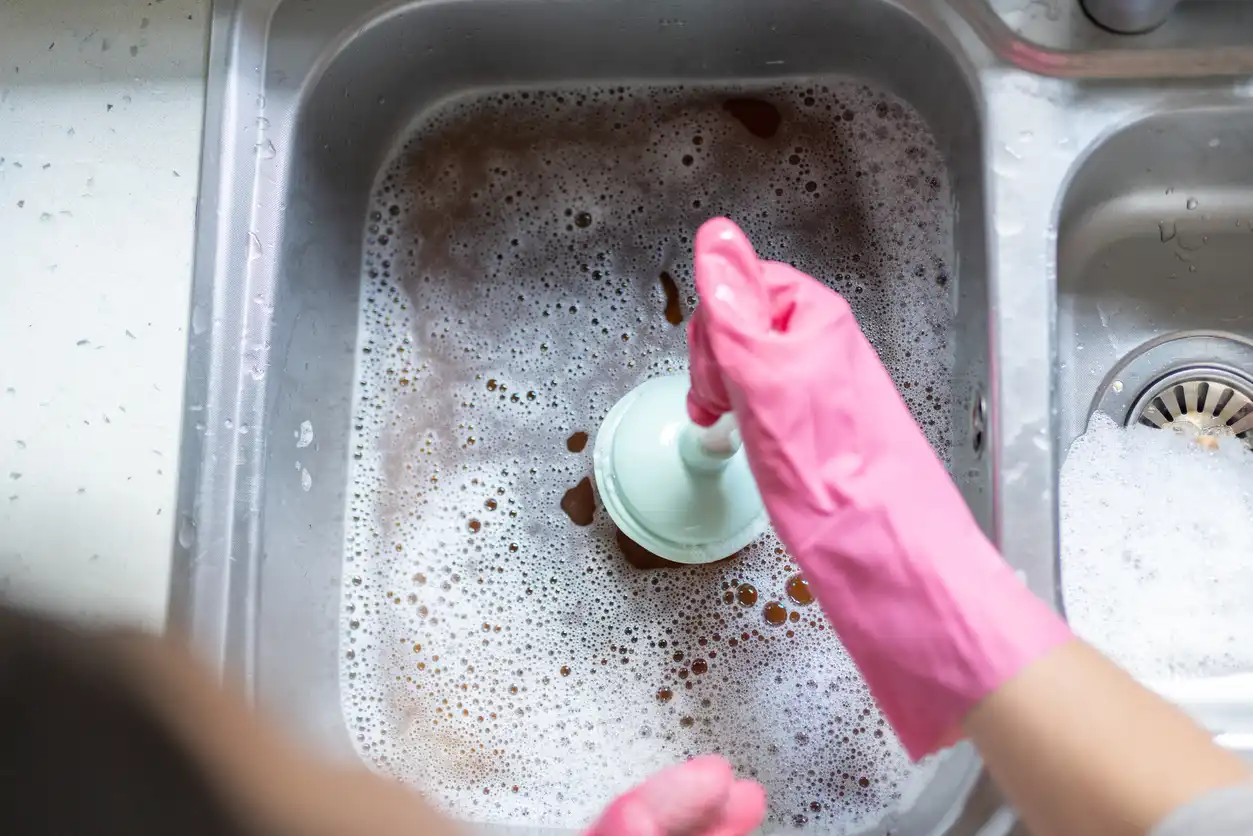 A close-up view of a person wearing pink gloves using a small teal plunger to unclog a kitchen sink