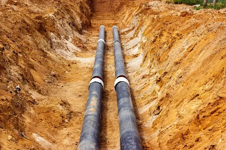 Two large, black pipes laid side-by-side in a deep trench of brown dirt. The trench is surrounded by more loose dirt